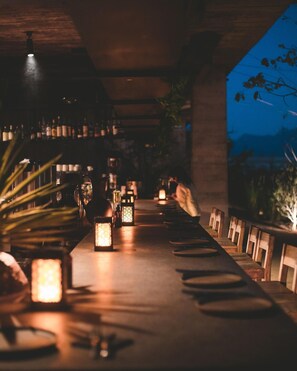 A dimly lit bar area with wooden tables and chairs, illuminated by lanterns, and a view of the outdoors. - Paradero Todos Santos (Todos Santos)