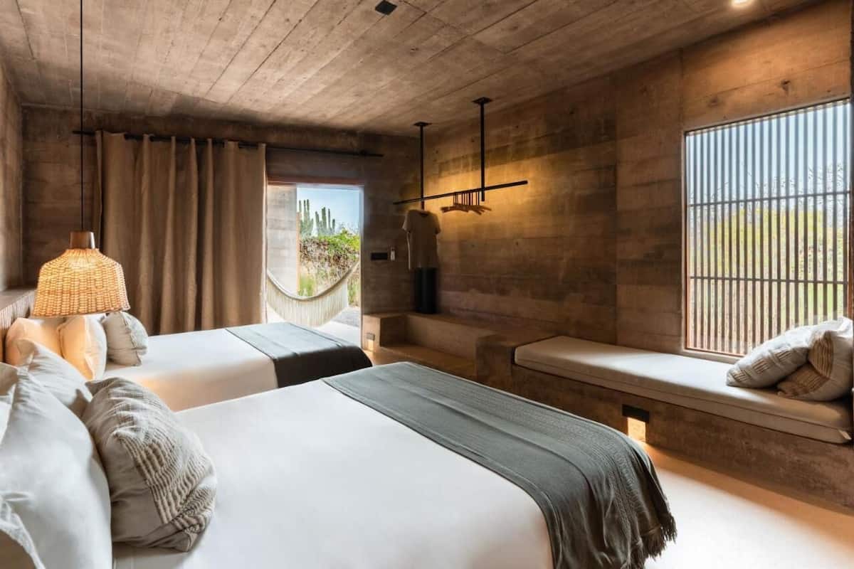 Quarto moderno com duas camas, um banco, uma luminária pendente e vista para o exterior.