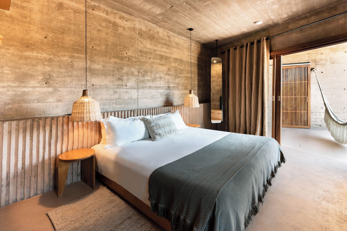 Quarto moderno com cama grande, cabeceira de madeira e vista para uma estrutura de madeira lá fora.