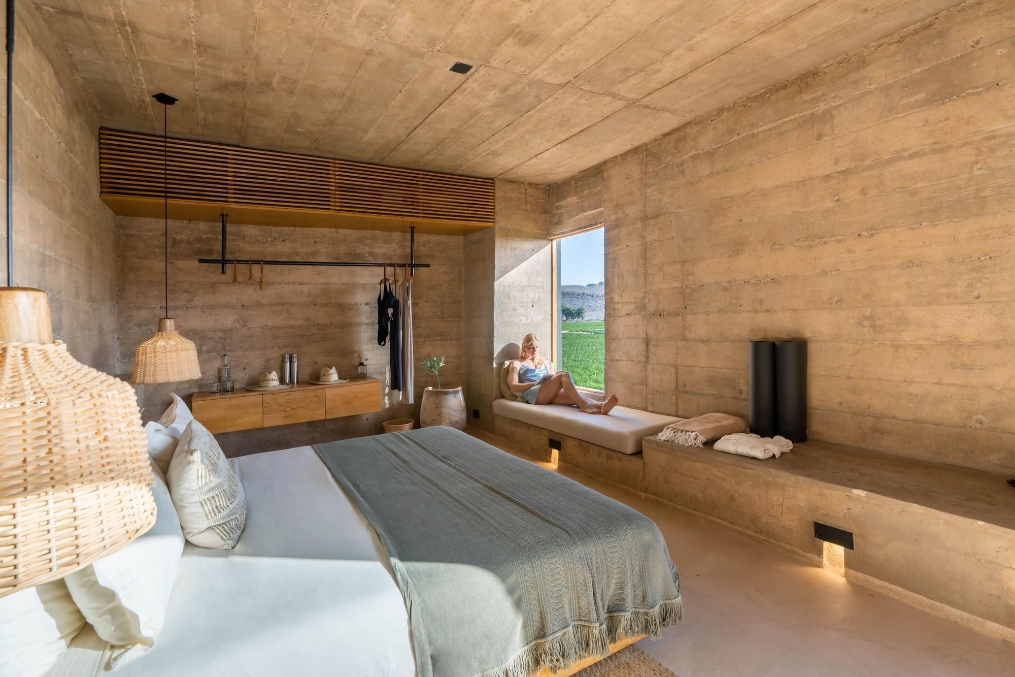Un dormitorio con cama, mesita de noche, lámparas colgantes, un banco con vistas y un ventanal grande.