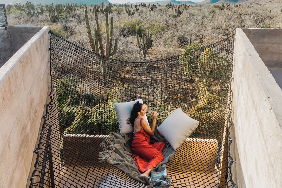 Uma pessoa sentada numa rede em paisagem desértica, com cactos e montanhas ao fundo.