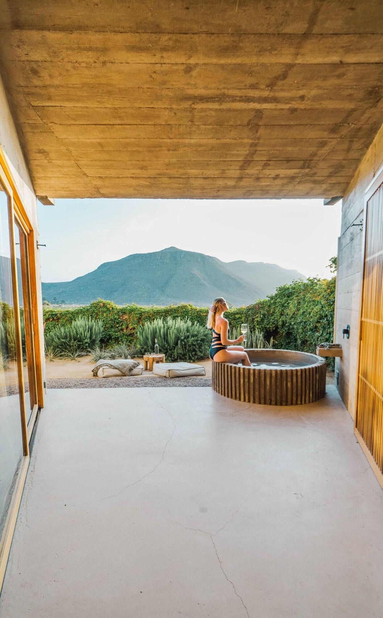 A covered outdoor area with a hot tub, a person relaxing, and a mountainous backdrop.