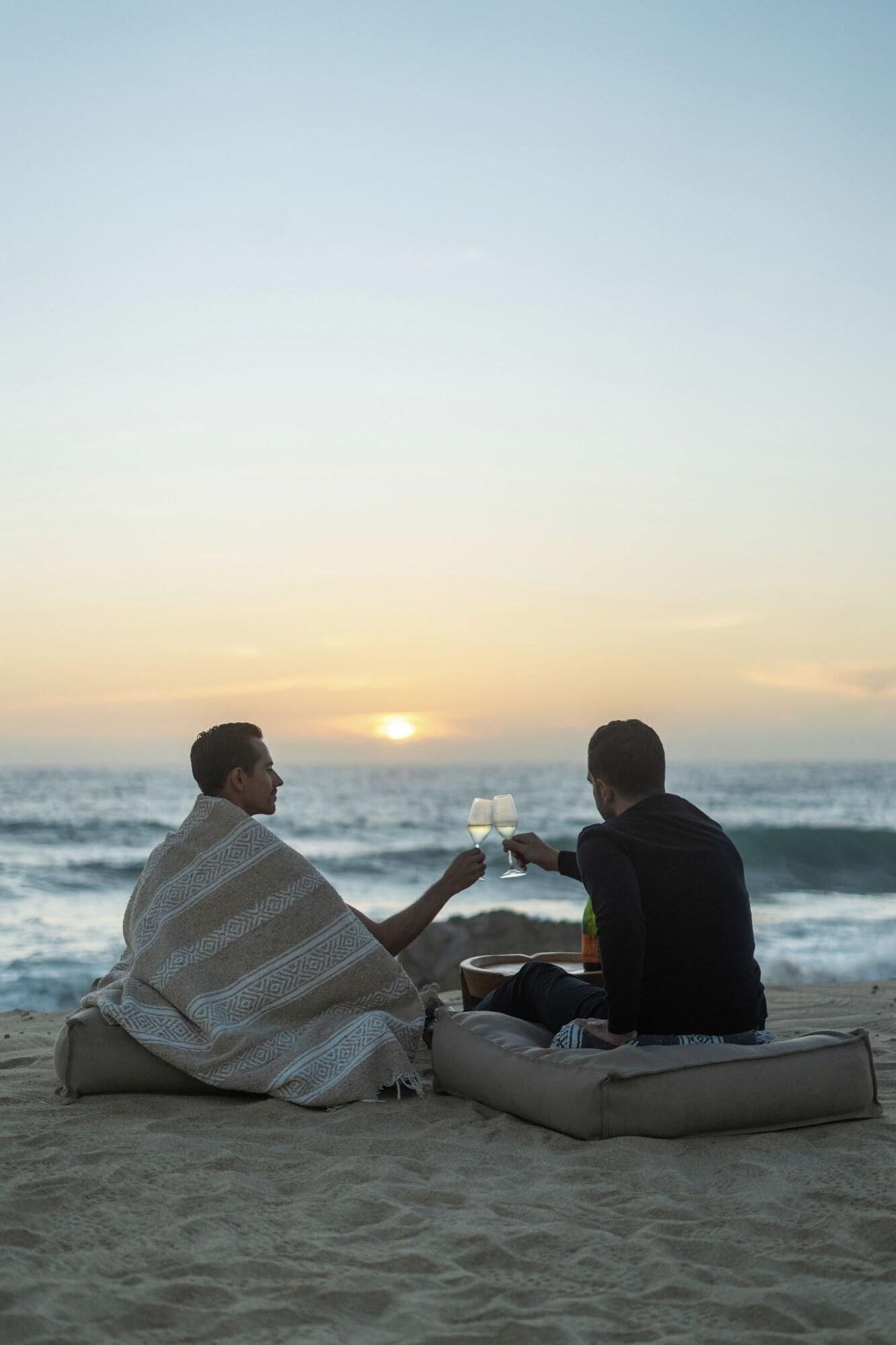 Two people sitting on a beach blanket facing the ocean, holding glasses.