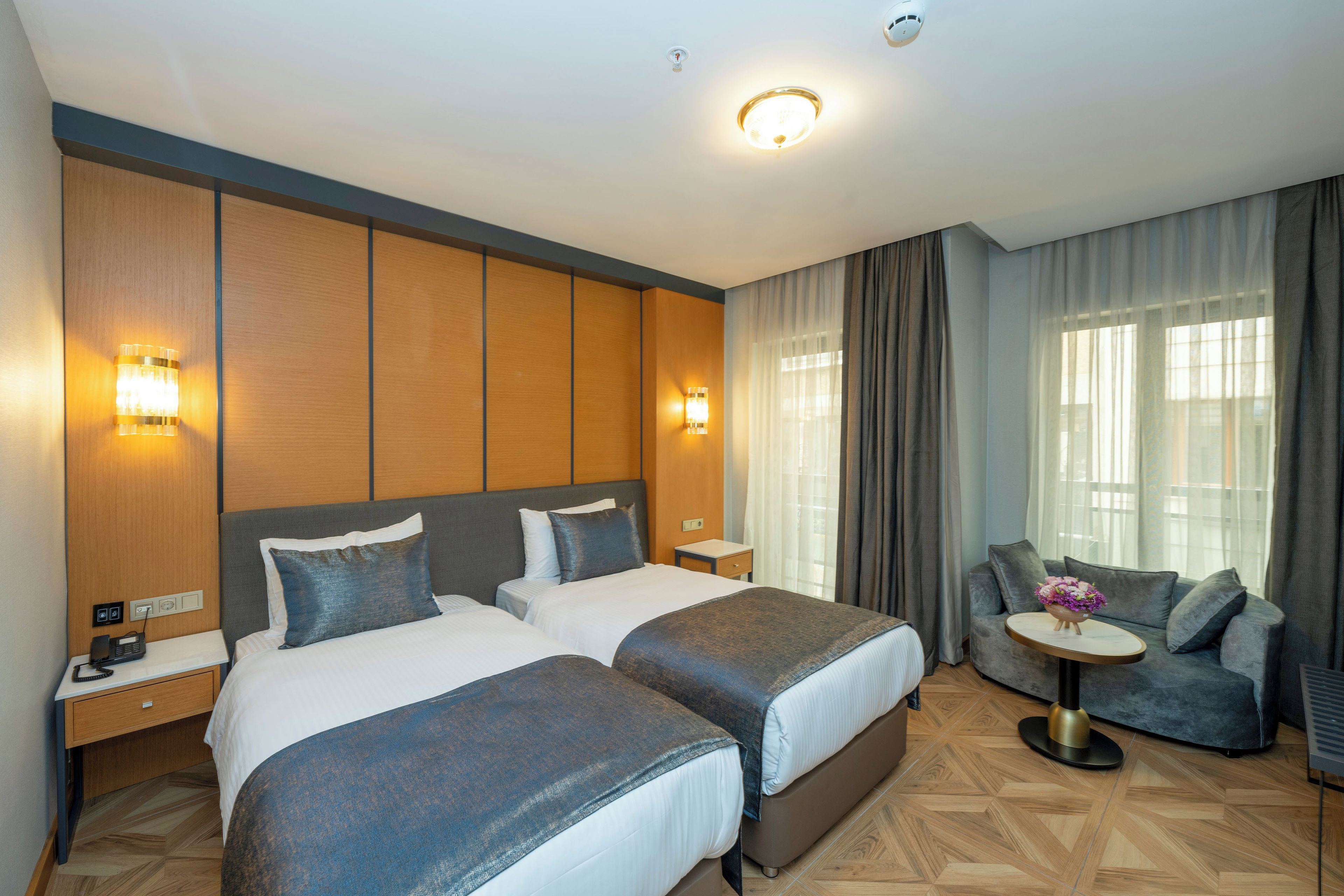 Quarto de hotel com duas camas, um sofá, uma mesa redonda pequena e vista da cidade através de grandes janelas.