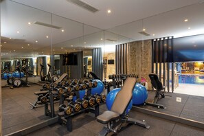 Un gimnasio con equipo de ejercicio, un espejo grande y una zona de piscina visible a través del vidrio.
