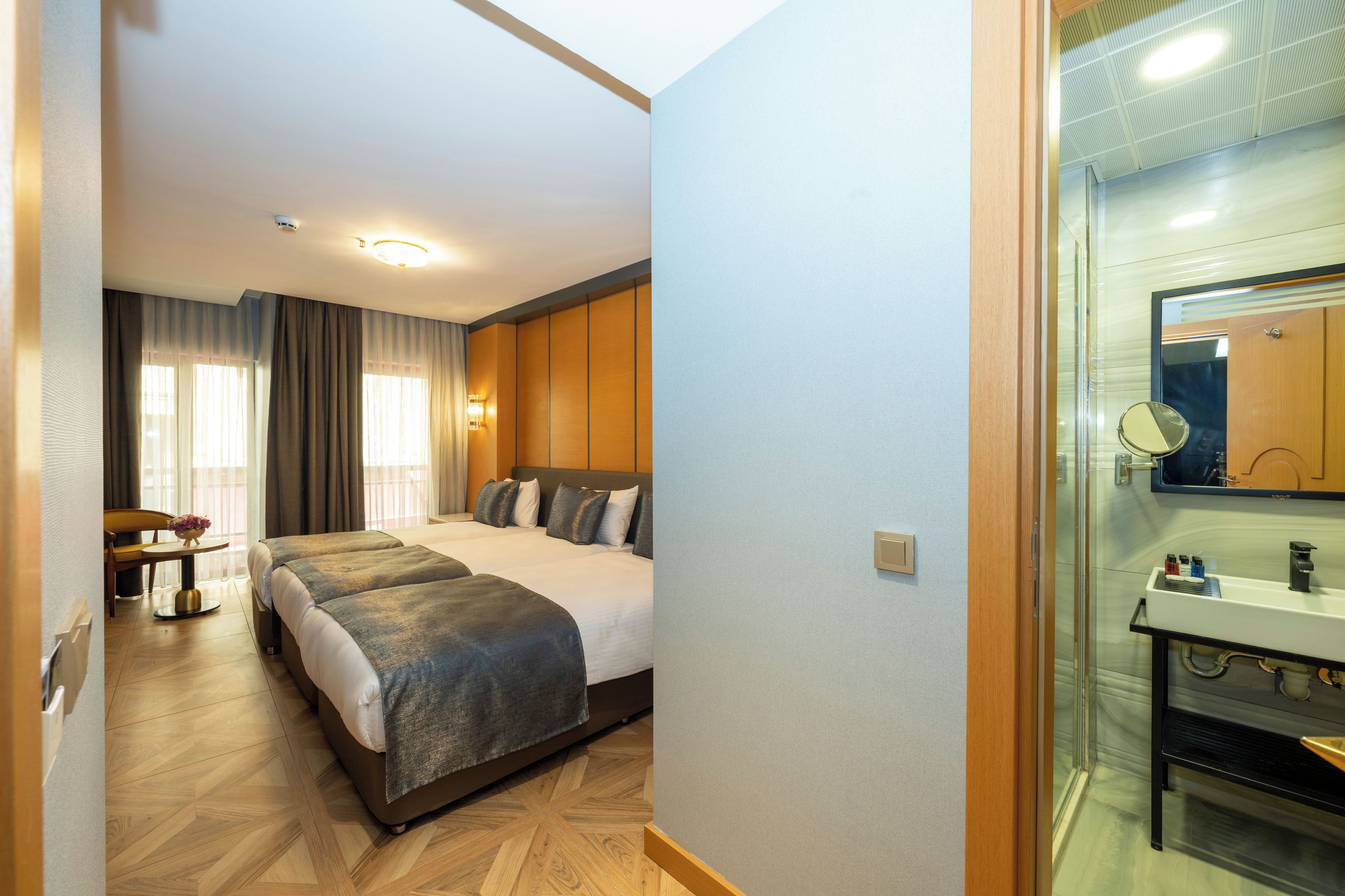 Habitación de hotel con cama doble, un escritorio con silla, una mesita con un florero y un baño con lavabo y espejo.
