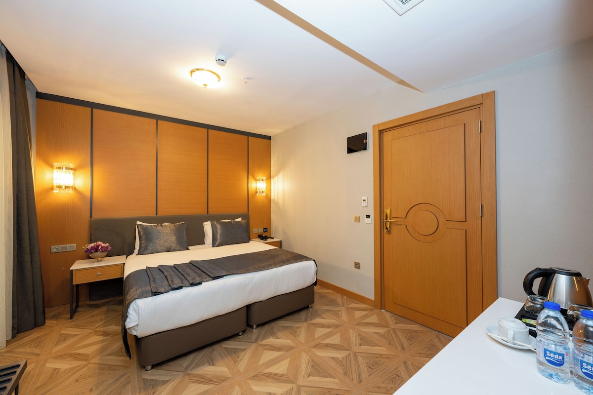 A hotel room with a large bed, wooden panel walls, a nightstand with a lamp, and a wooden door.