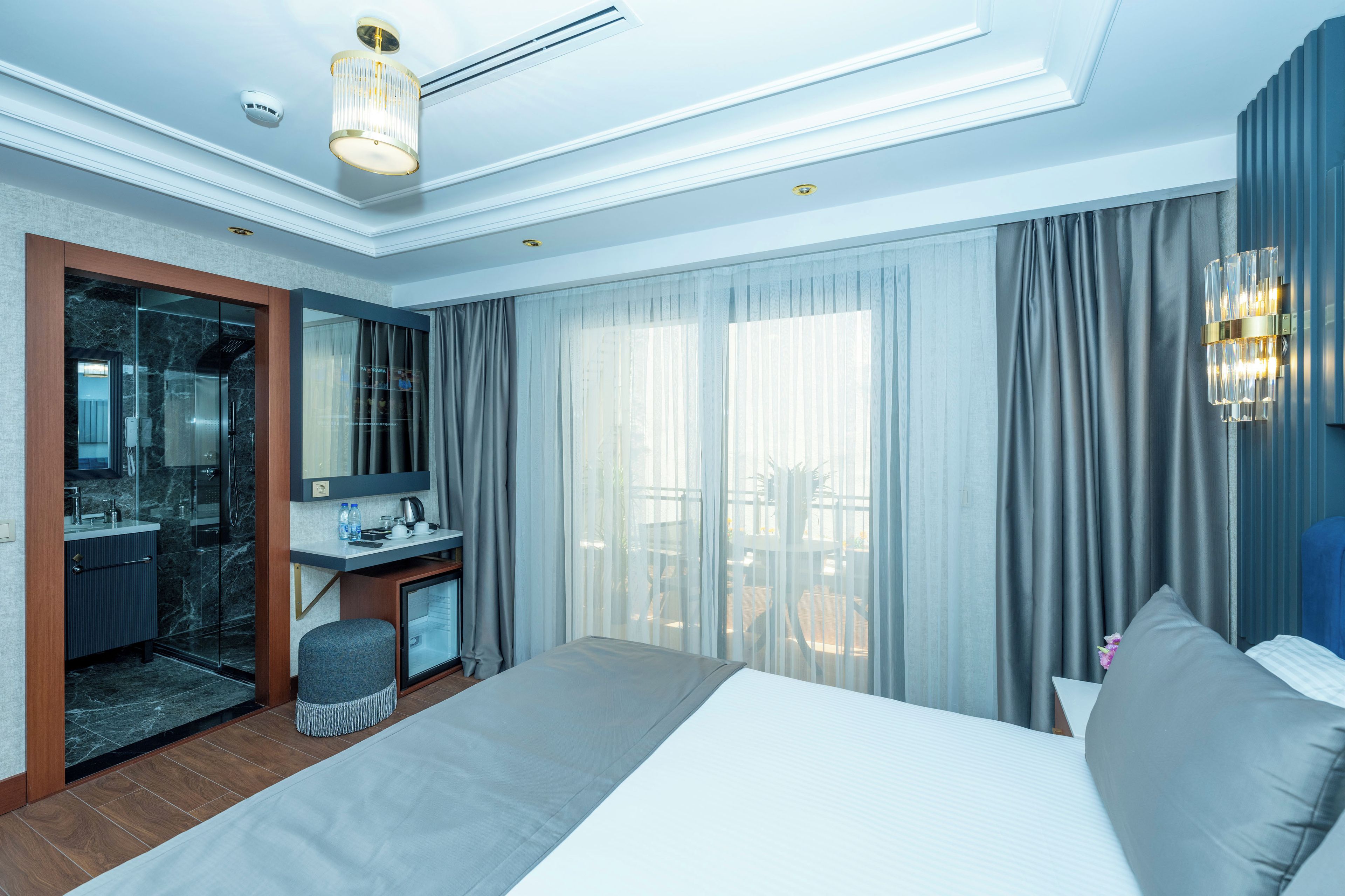 Ein modernes Hotelzimmer mit einem großen Bett, einem Schreibtisch mit Stuhl, einer Duschkabine aus Glas und einem Balkon mit Vorhängen.