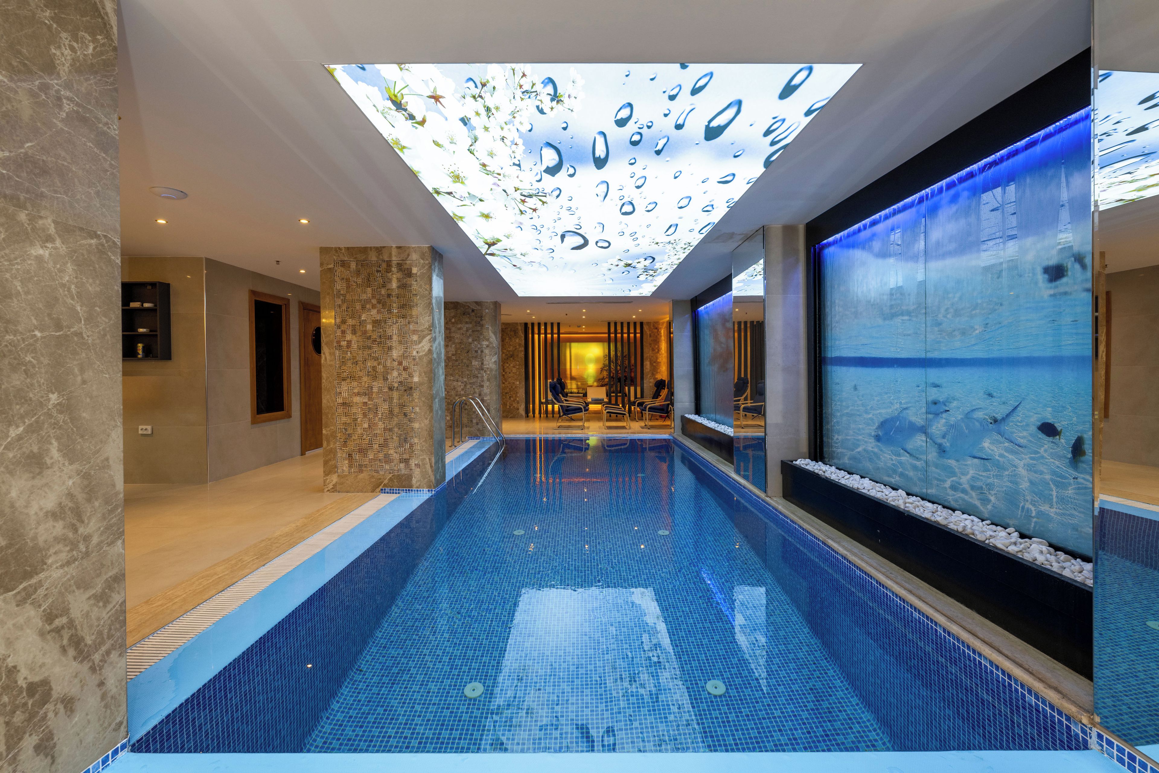 Una piscina interior moderna con un techo que imita una superficie acuática, un área de descanso con sillones y vistas a la piscina.
