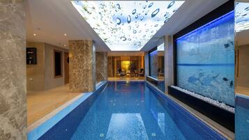 Una piscina interior moderna con un techo que imita una superficie acuática, un área de descanso con sillones y vistas a la piscina.