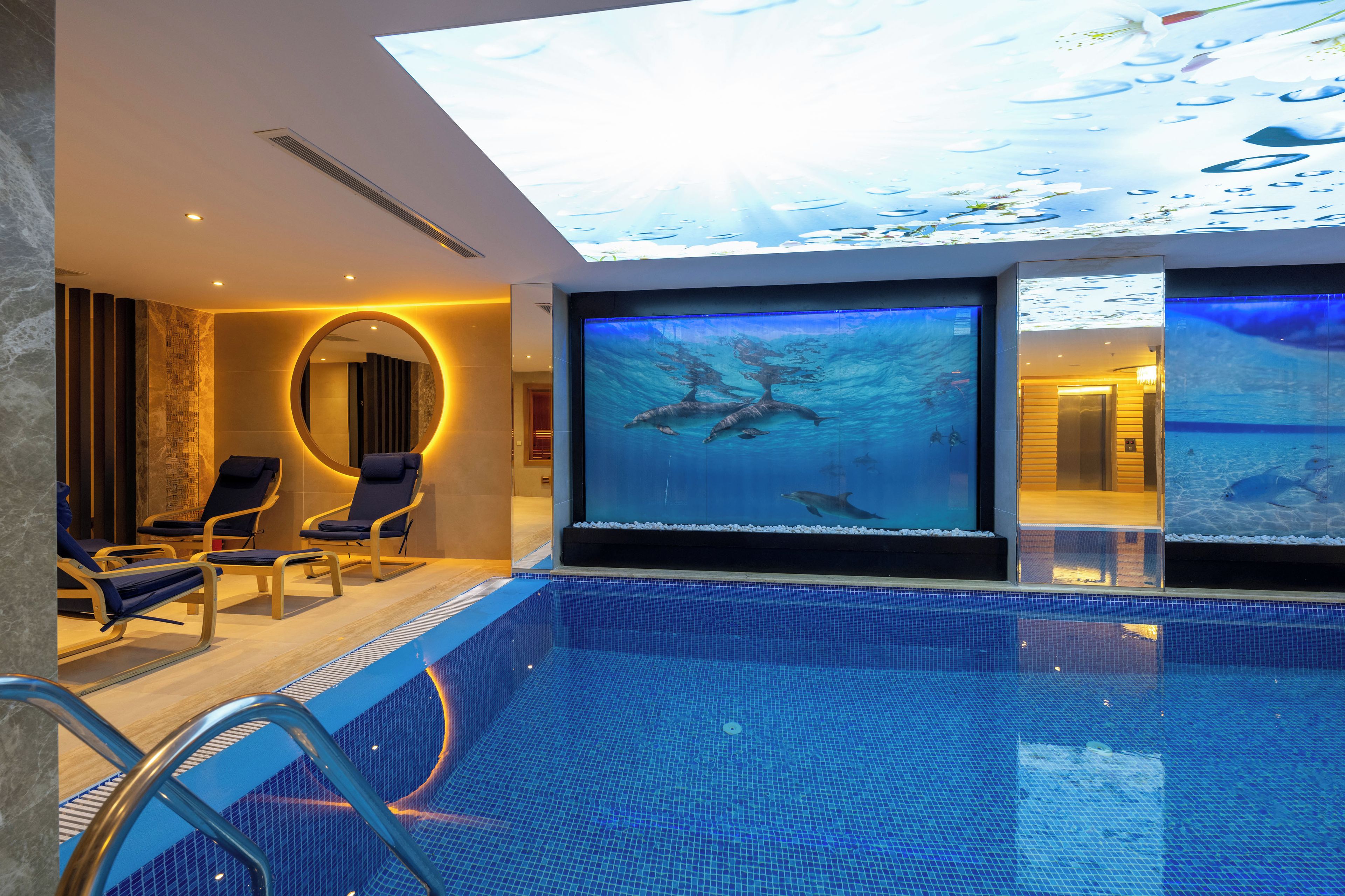 Una piscina interior moderna con un gran acuario, sillas de descanso y un tragaluz.