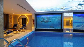 Una piscina interior moderna con un gran acuario, sillas de descanso y un tragaluz.