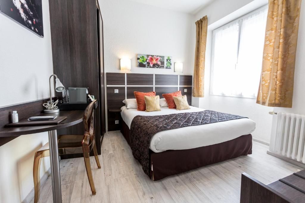 A modern hotel room with a bed, bedside table, desk, chair, and a window with curtains.