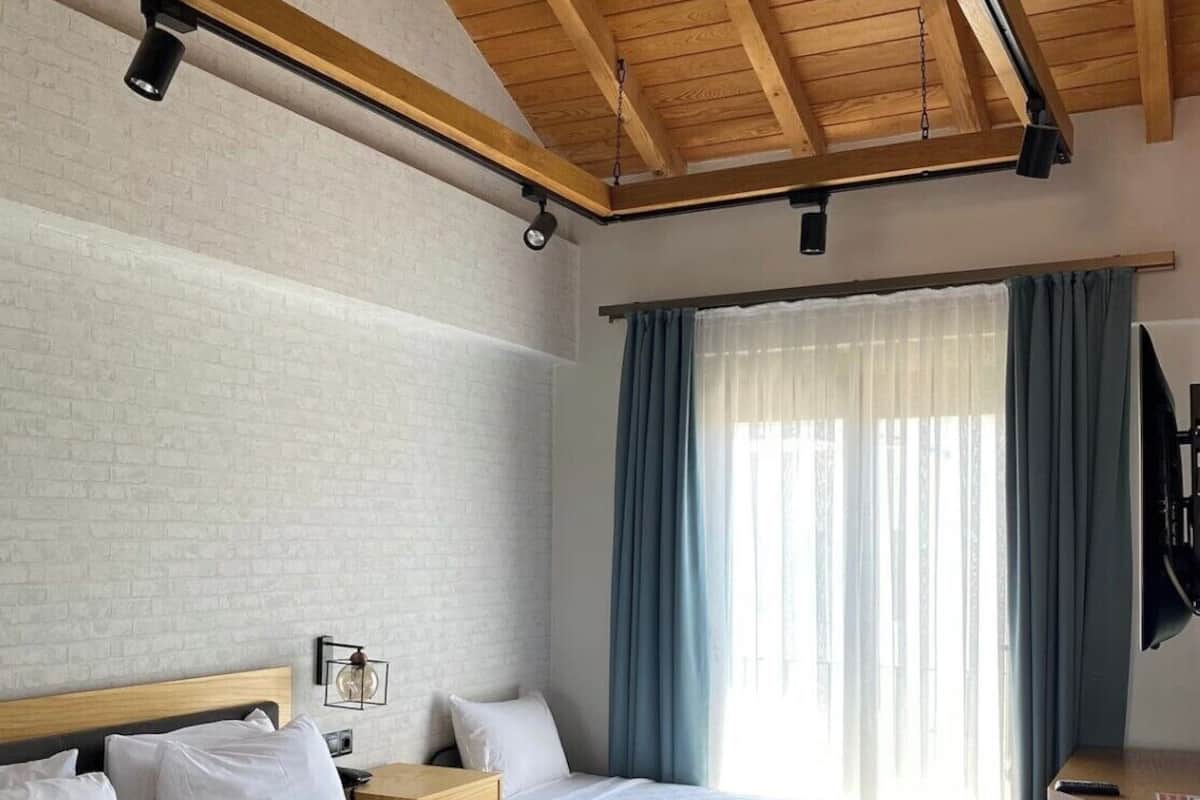 A hotel room with two beds, a wooden ceiling, a brick wall, a window with curtains, and a flat-screen TV.