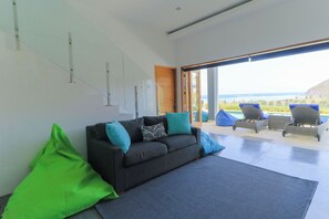 Ein modernes Wohnzimmer mit einer Couch, Kissen, einem großen Fenster und Blick ins Freie.