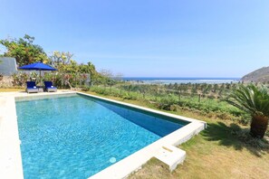 Uma piscina retangular com água azul cristalina, rodeada por uma área gramada com vista para o oceano e colinas ao fundo.