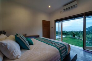A bedroom with a bed, pillows, a bedside table, a nightstand, an air conditioning unit, and a sliding glass door leading to a balcony with a view of the landscape. - Villa K by Zuzu (Selong Belanak)