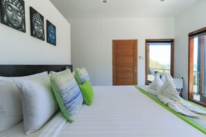 Quarto de hotel com cama, almofadas, obra de arte na parede, porta de madeira e vista pela janela.