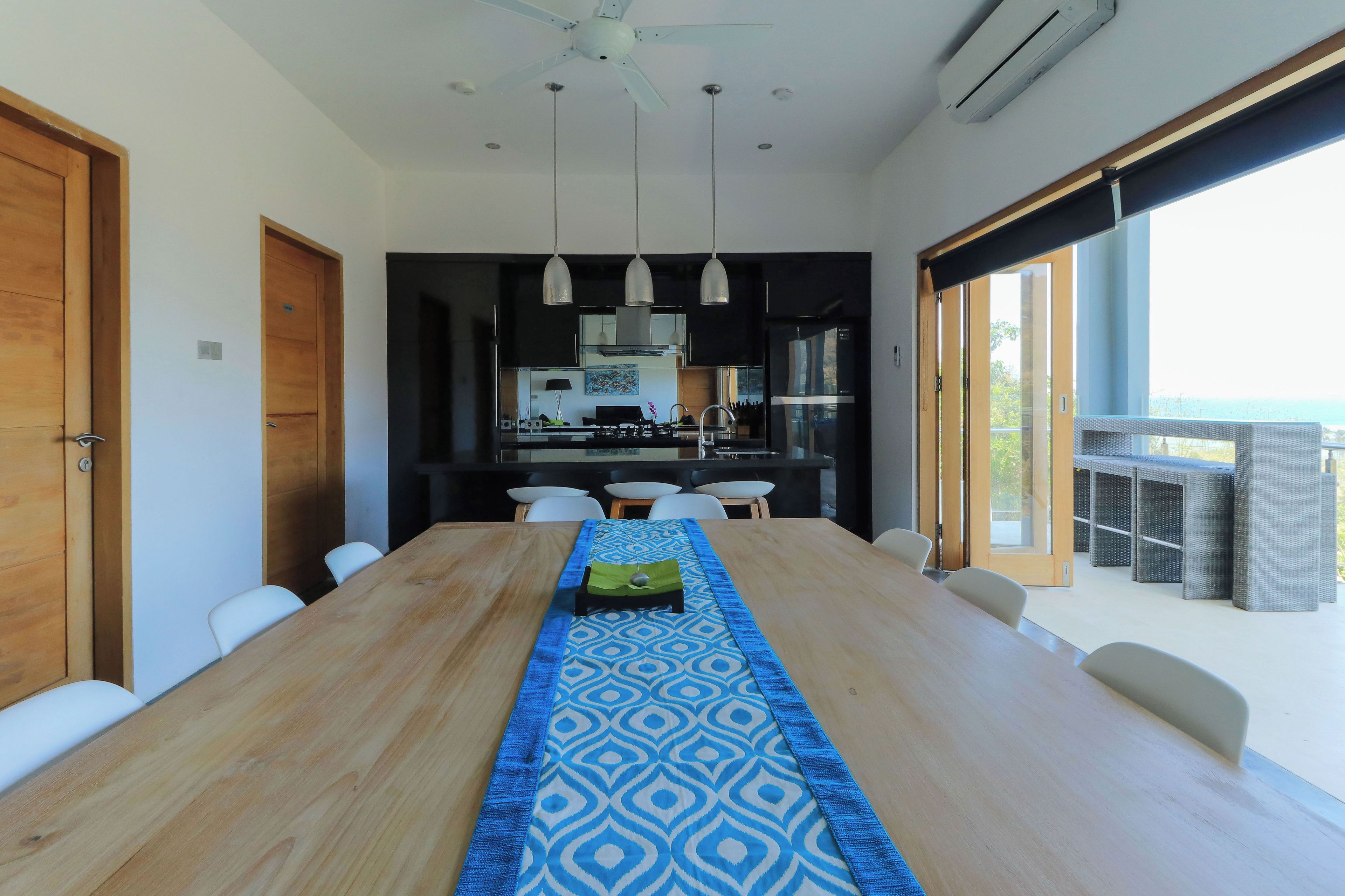 Uma área de jantar moderna com uma mesa de madeira comprida, luminárias pendentes e vista para o exterior.