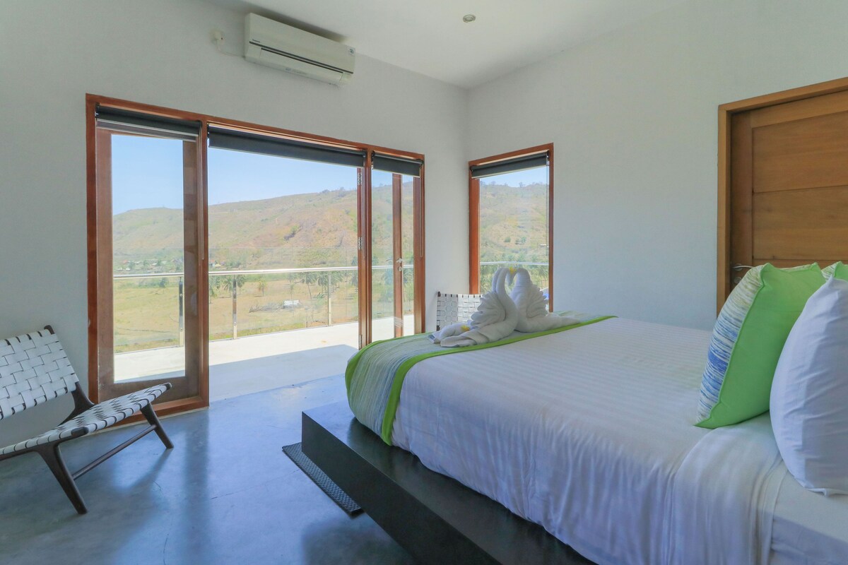 A bedroom with a bed, a bench, and large windows with a view of hills.