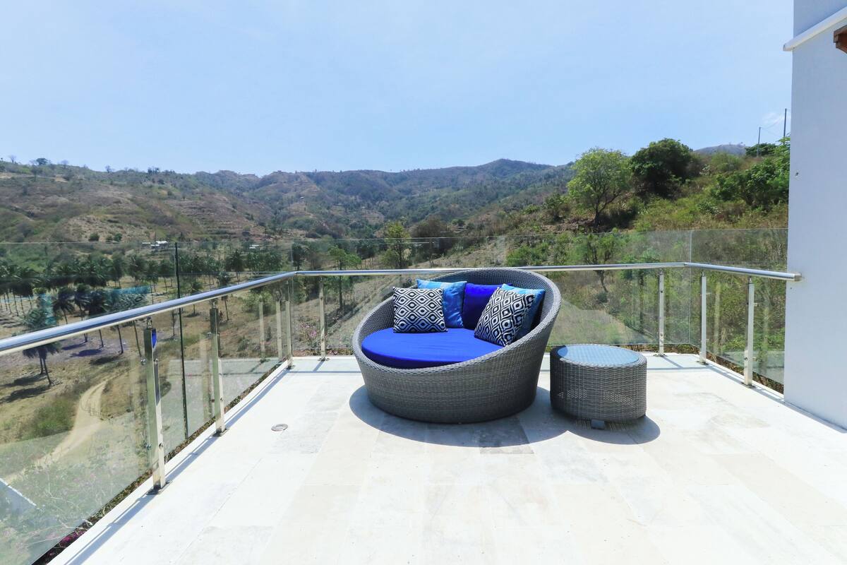 Un balcón con un sofá y reposapiés de mimbre, con vistas a un paisaje montañoso.