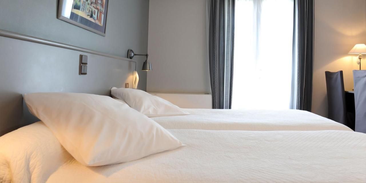 Quarto de hotel com duas camas, uma mesa de cabeceira, uma luminária e uma janela com cortinas.