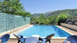 A rectangular swimming pool with clear blue water, surrounded by a low brick wall. There are several lounge chairs and a table near the pool. The area is enclosed by a green fence, and the background features a hilly landscape. - Logis Hotel Du Fronton-agian (Itxassou)