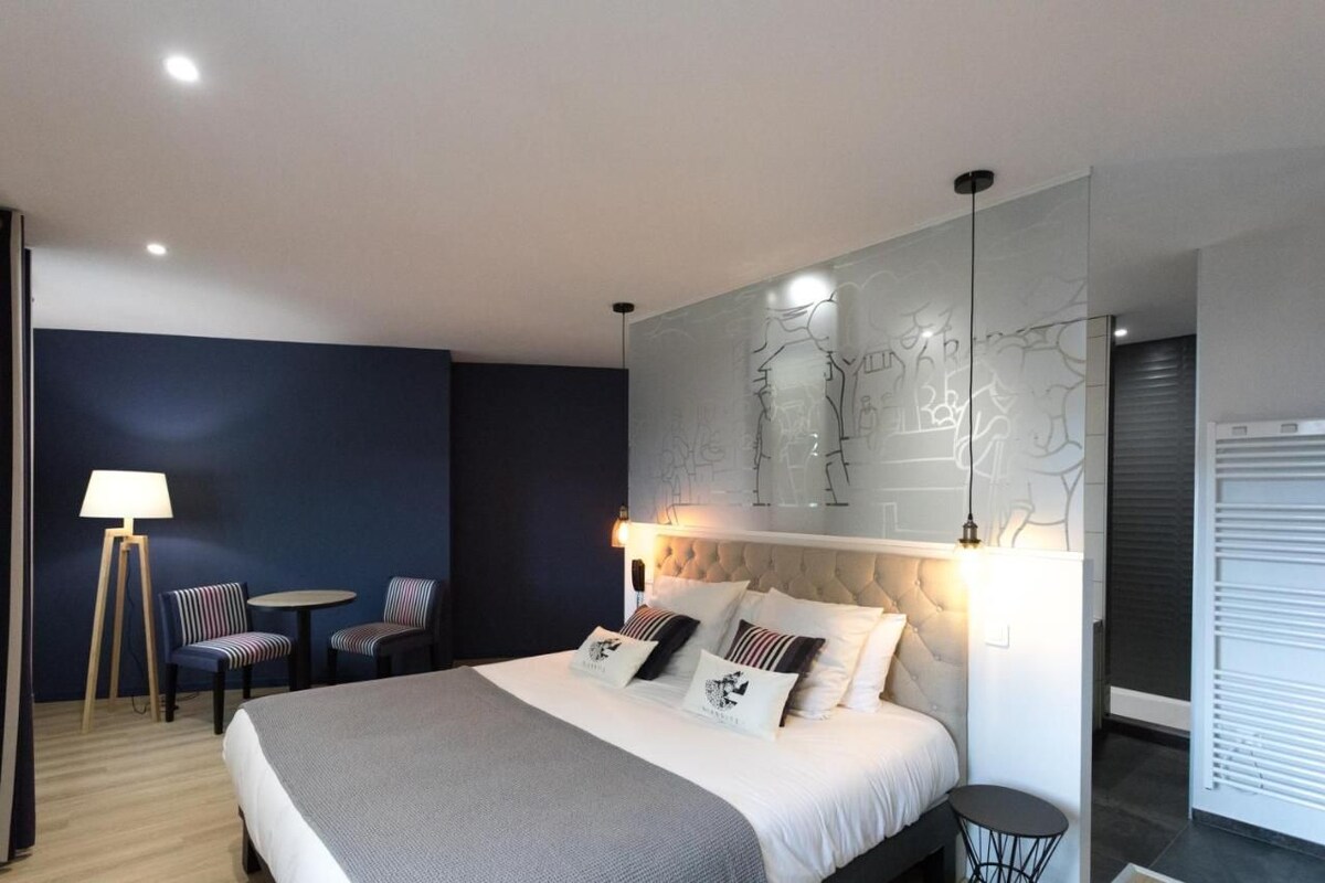 A modern hotel room with a large bed, a wall-mounted artwork, a small round table, two chairs, and a lamp.
