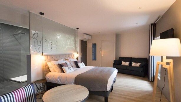 A modern hotel room with a large bed, a sofa, a small round table, and a standing lamp. - Logis Hotel Du Fronton-agian (Itxassou)