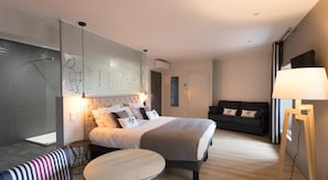 A modern hotel room with a large bed, a sofa, a small round table, and a standing lamp. - Logis Hotel Du Fronton-agian (Itxassou)