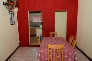 Une pièce de taille modeste comprenant une table à manger, des chaises, un ventilateur et un coin cuisine visible à travers une porte ouverte.