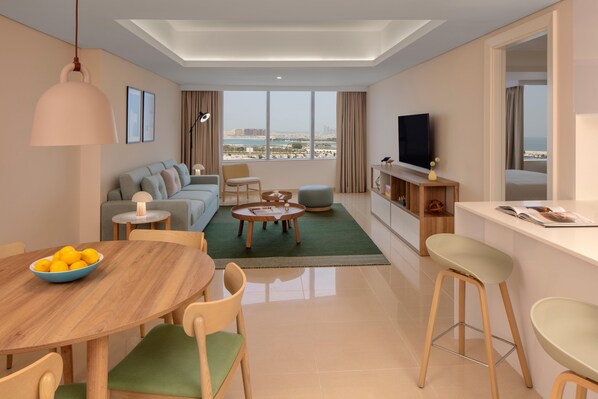 A modern living room with a sofa, coffee table, and TV, a dining area with a table and chairs, and a view of the city through large windows. - Staybridge Suites Dubai Internet City (DUBAI)