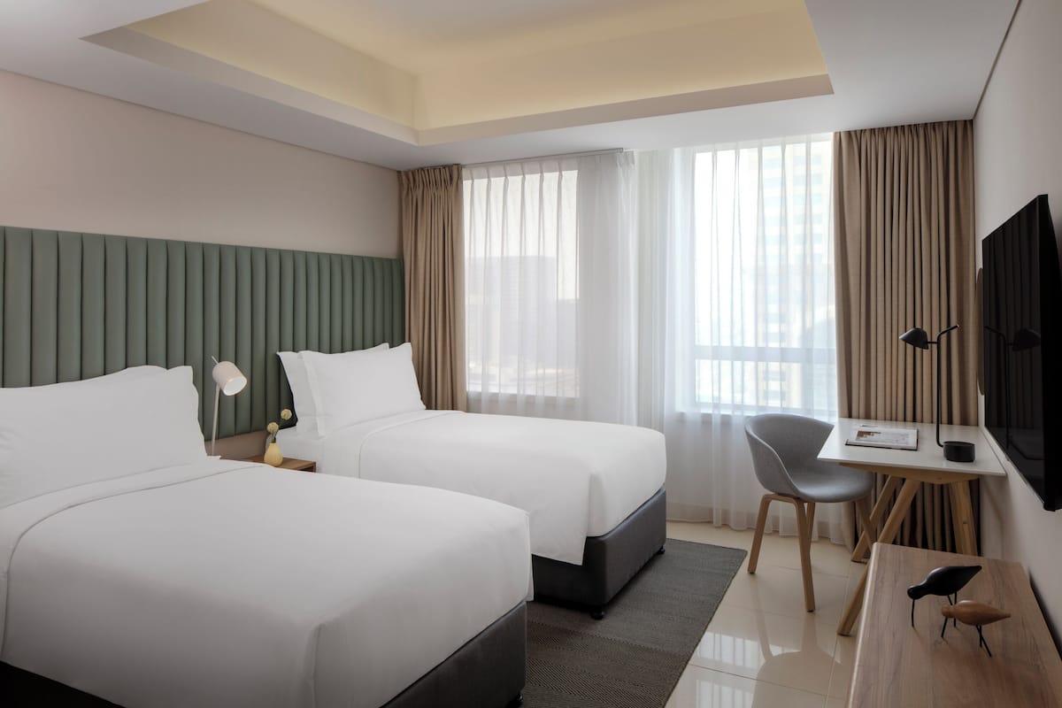 Ein Hotelzimmer mit zwei Betten, einem Schreibtisch, einem Sessel, einem Fernseher und einem Fenster mit Vorhängen.