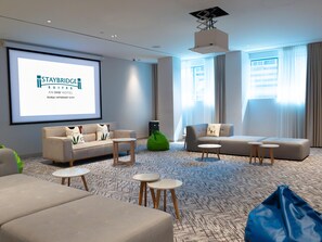 A hotel room with a large screen displaying Staybridge Suites, a sofa, a coffee table, a bean bag, and a view of the city through large windows. - Staybridge Suites Dubai Internet City (DUBAI)