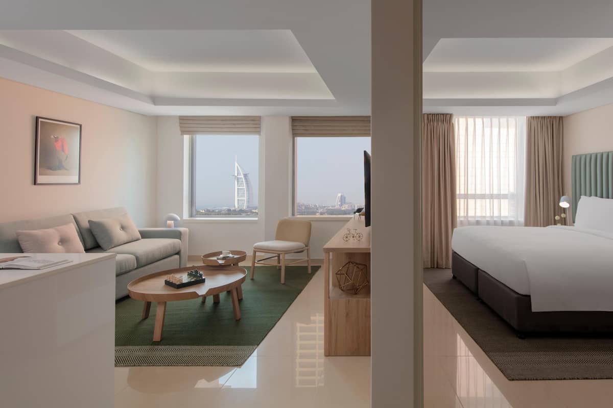Ein modernes Hotelzimmer mit einem Bett, einer Couch, einem Schreibtisch und einem Sessel. Es gibt Fenster mit Blick auf eine Stadtansicht und ein Gebäude.