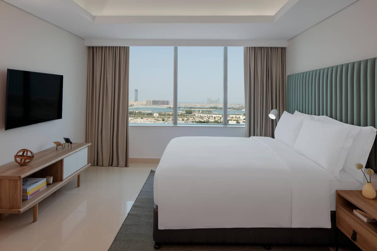 Ein modernes Hotelzimmer mit einem großen Bett, einem Flachbildfernseher, einem Holztischchen und Blick auf eine Stadtlandschaft durch große Fenster.