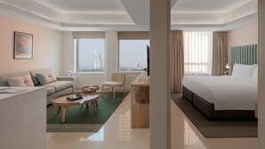 A modern hotel room with a bed, a sofa, a desk, and a chair. There are windows with a view of a cityscape and a building.