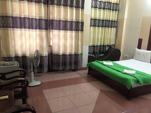 Ein Hotelzimmer mit Bett, Sessel, Schreibtisch, Ventilator und Vorhängen.