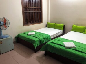 Une chambre avec deux lits, chacun agrémenté de draps verts et de draps blancs, une fenêtre avec des barreaux, un ventilateur sur pied et une petite table sur laquelle se trouve un objet bleu.