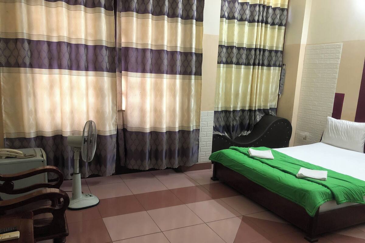 Ein Hotelzimmer mit Bett, Sessel, Schreibtisch, Ventilator und Vorhängen.