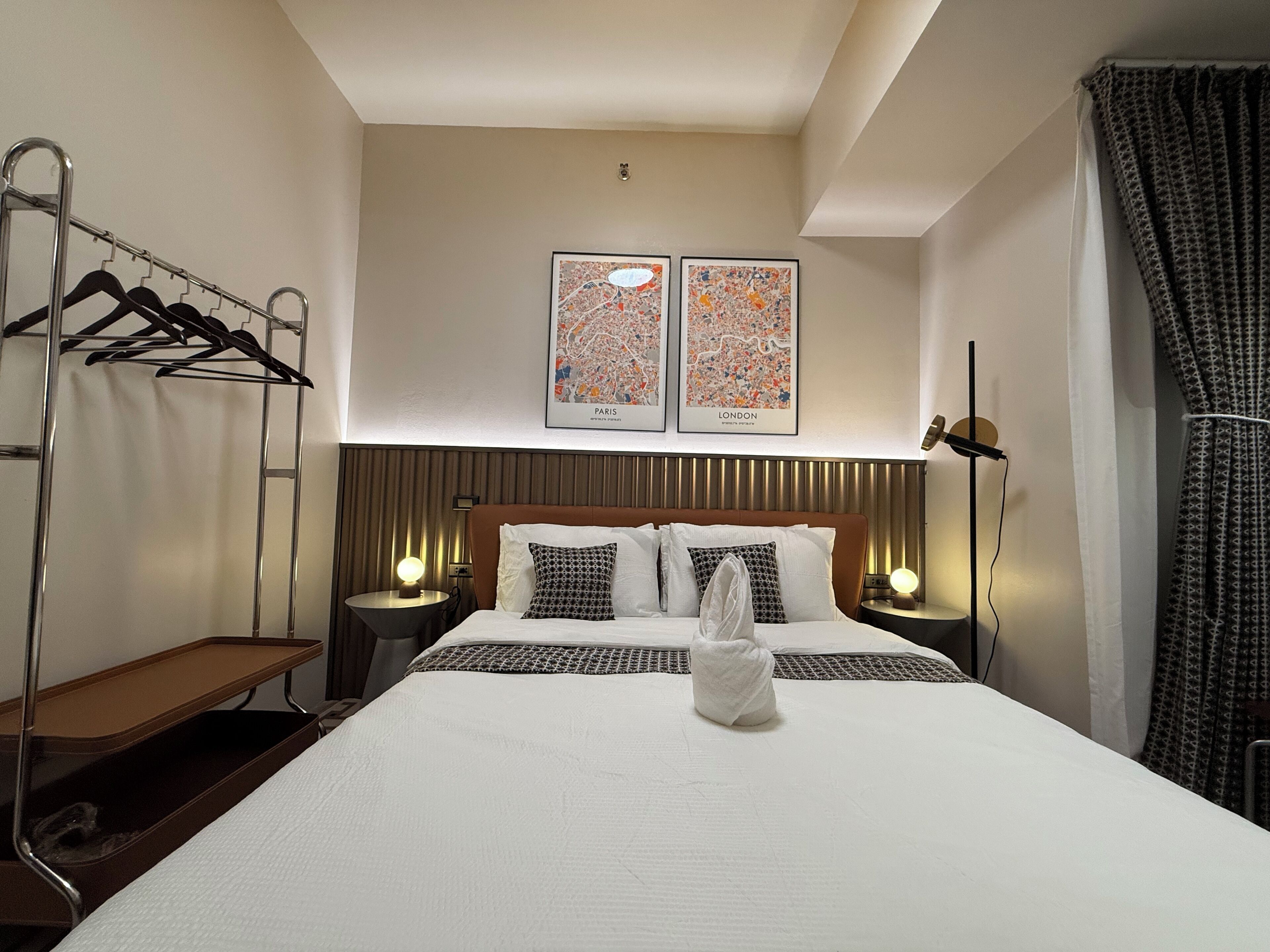 A hotel room with a bed, two wall-mounted lamps, a coat rack, and two framed cityscape pictures.
