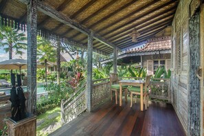 A covered outdoor area with a wooden table and chairs, surrounded by lush greenery and a view of a pool.