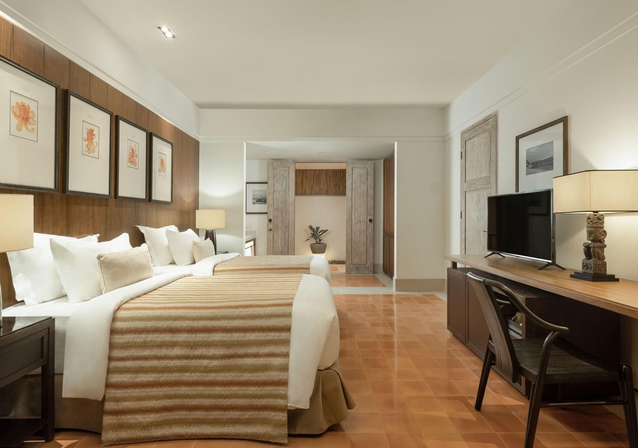 A hotel room with two beds, a desk with a television, and framed artwork on the walls.