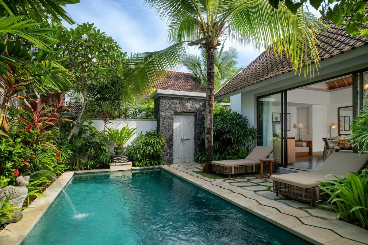 Una piscina con sillones de descanso, rodeada de plantas tropicales y un muro de piedra.