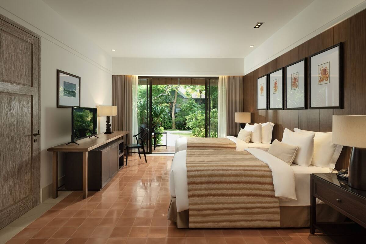Habitación de hotel con cama, escritorio, televisor y ventanas amplias con vistas a zonas verdes.