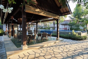 An outdoor patio with a pool, covered seating area, and stone flooring.
