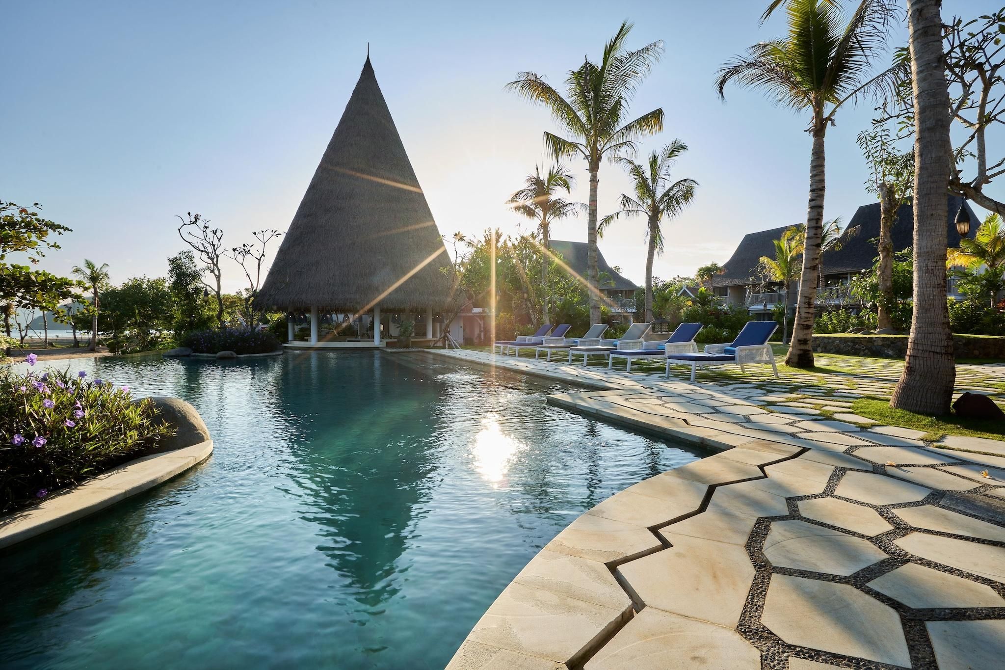 Una piscina con sillones de descanso, palmeras y una estructura de techo de paja.