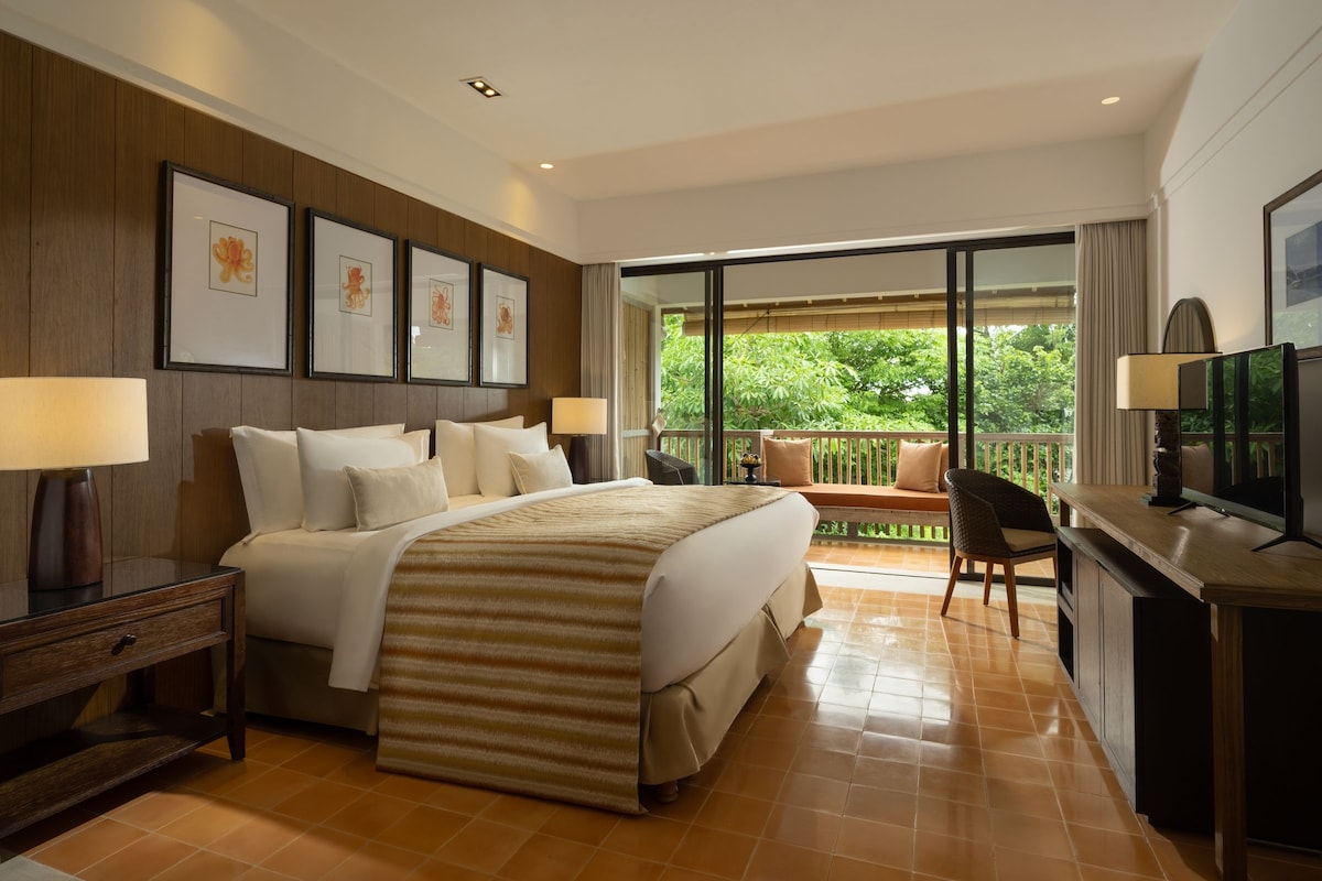 A hotel room with a bed, bedside lamps, a desk with a television, a chair, and a balcony with a view of greenery.