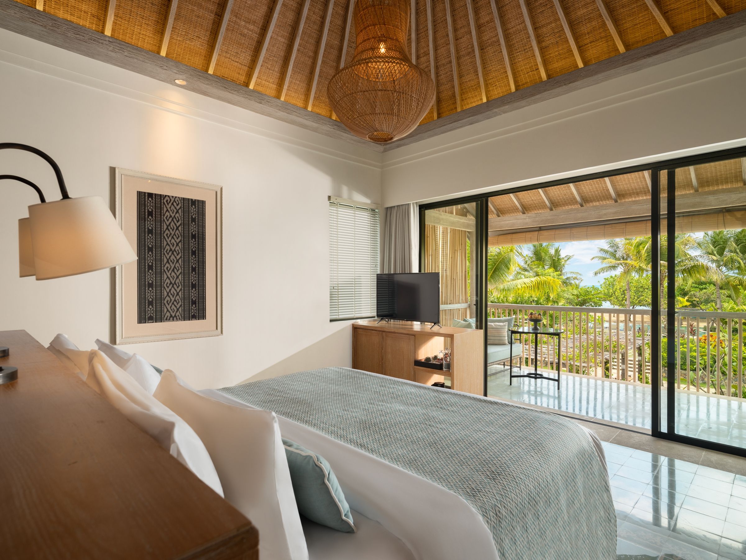 Un dormitorio con cama, televisor, escritorio y un ventanal con vistas a palmeras.