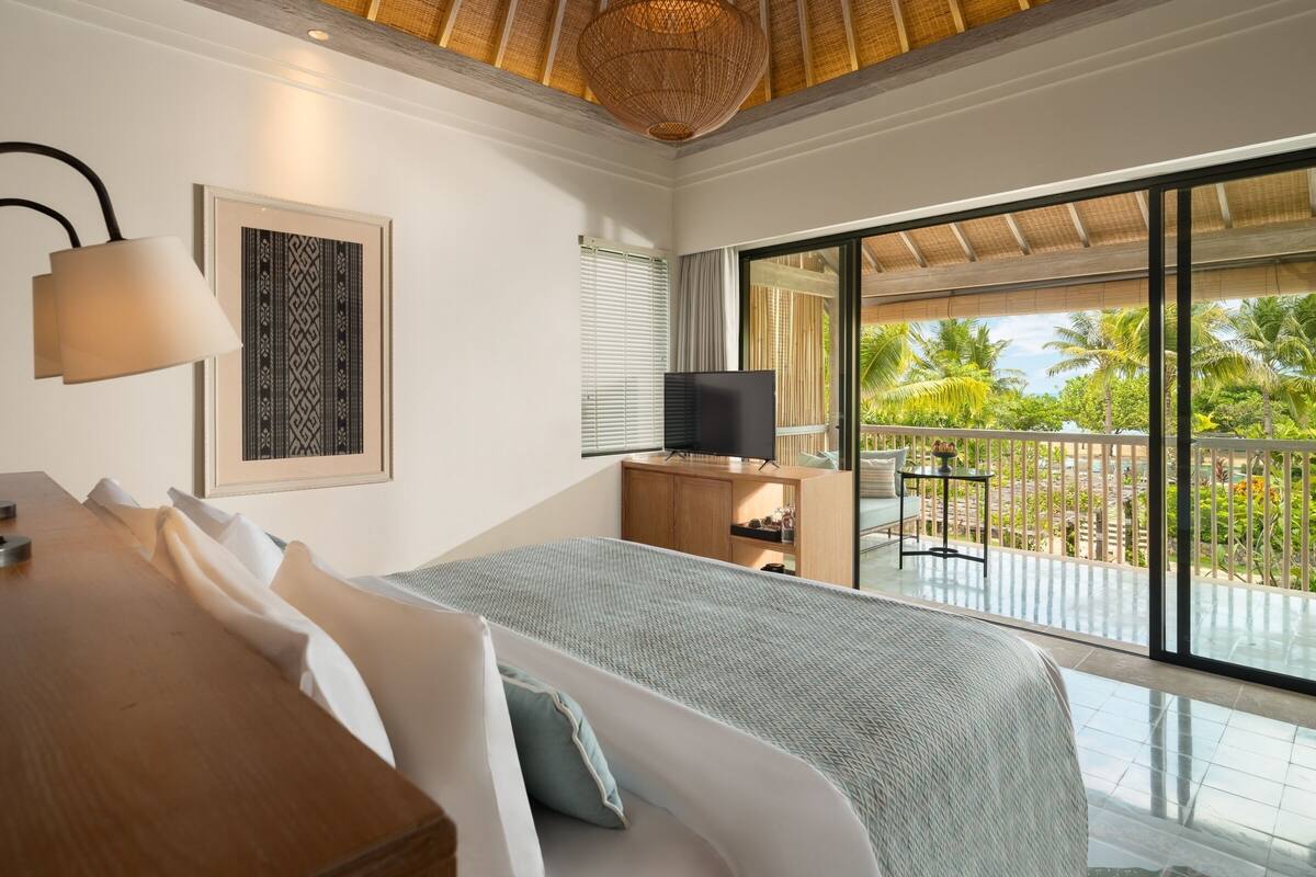 Un dormitorio con cama, televisor, escritorio y un ventanal con vistas a palmeras.